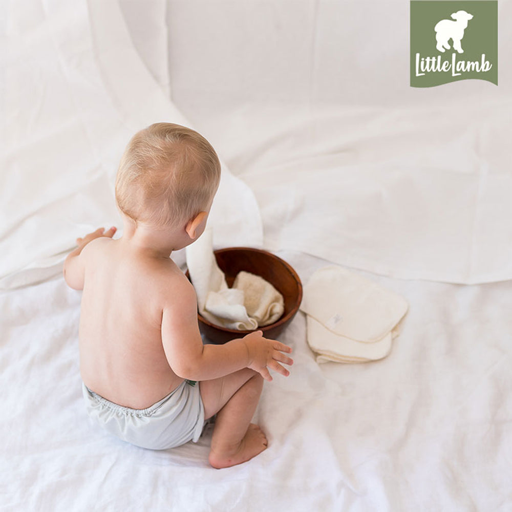 Baby sitzt mit einer Holzschale und einem Little Lamb Waschlappen auf einer weißen Decke