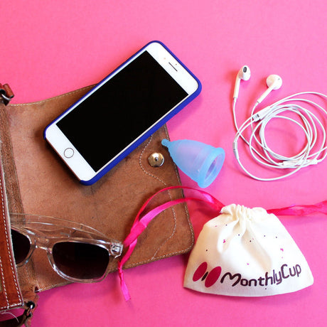 Stimmungsbild: MonthlyCup Menstruationstasse und Beutel in Handtasche mit Handy, Sonnenbrille und Kopfhörern