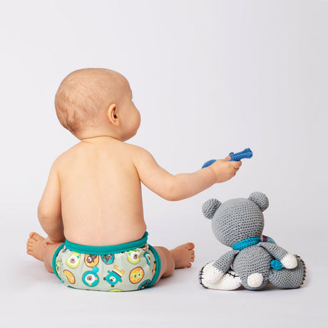 Baby sitzt mit Petit Lulu SIO-Überhose im Design Flying Bears neben einem Strickteddy, Ansicht von hinten