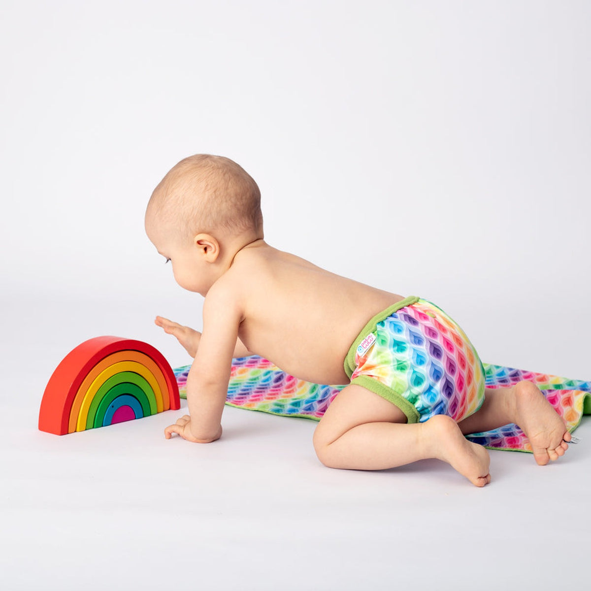 Krabbelndes Baby mit Petit Lulu SIO-Überhose im Design Rainbow Flames vor einem Holzregenbogen