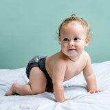 Lachendes Baby krabbelt auf einem Bett und trägt die Popolini EasyFree Wool in Anthrazit
